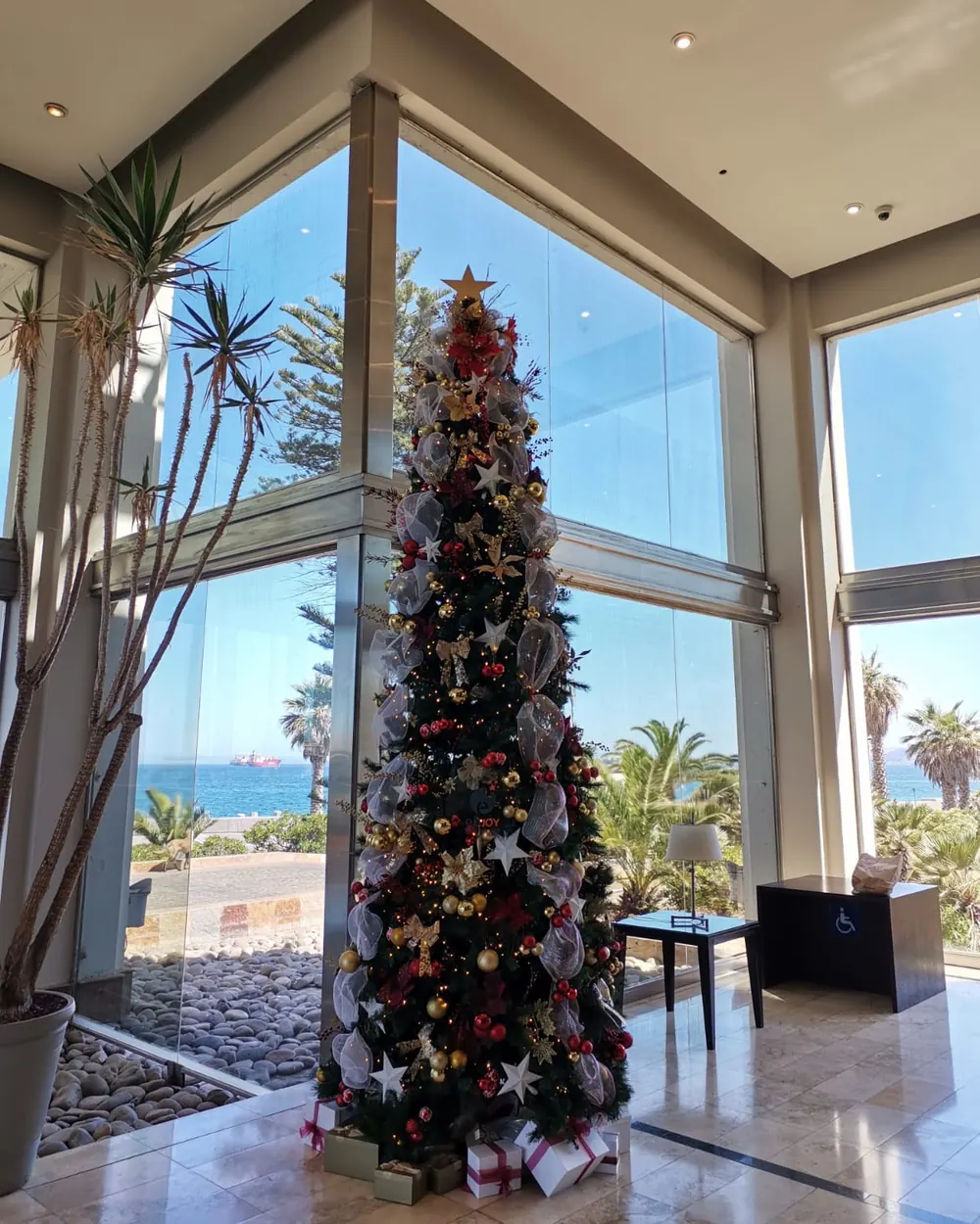 Árbol de Navidad de gran altura en rojo, dorado y plata, en un lobby con ventanales y vista al mar