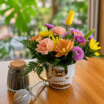 Taza blanca con diseños dorados rellena de flores pastel, frasco de té e infusor