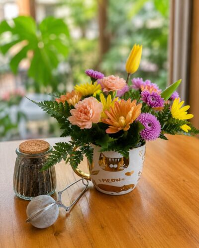 Taza blanca con diseños dorados rellena de flores pastel, frasco de té e infusor